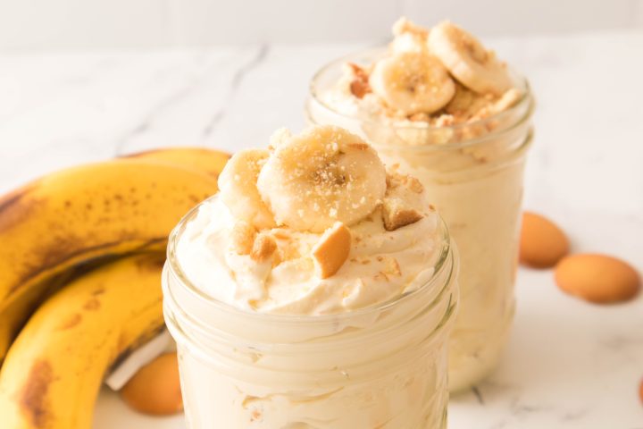 Banana pudding in a jar with banana slices and crushed vanilla wafers on top with another jar and bananas behind it 