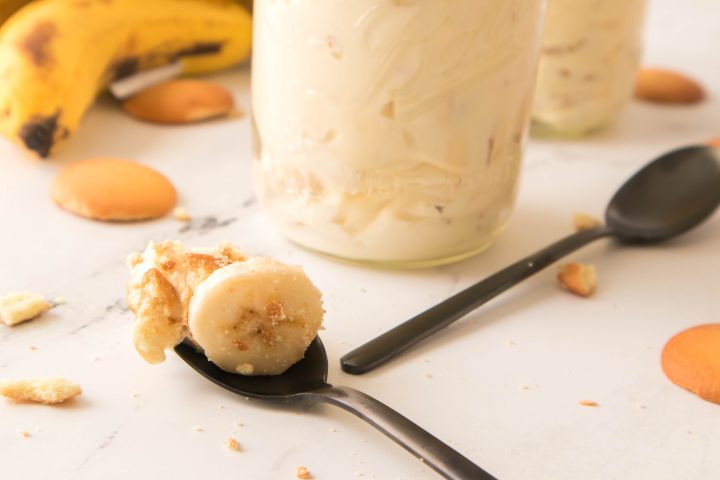 Picture of a banana slice with vanilla pudding on a spoon with a jar of banana pudding behind it 